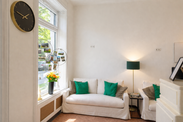 Cozy living room with white sofas, green pillows, a floor lamp, wall clock, and window with flowers.