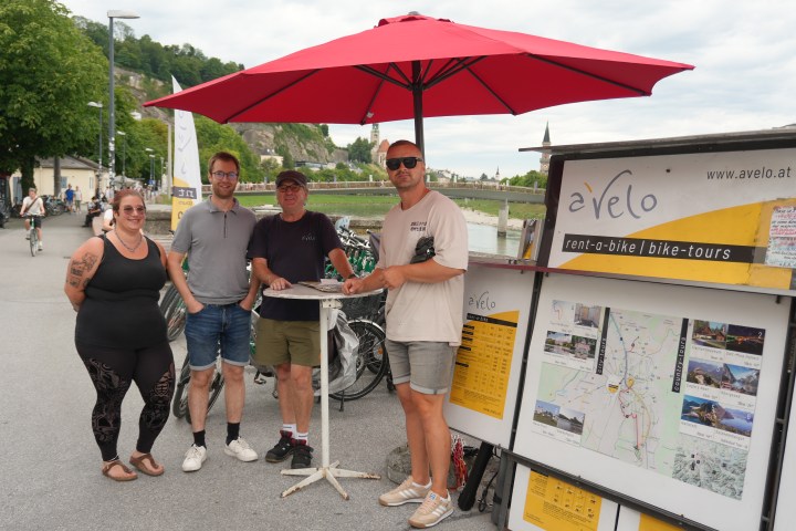 Four people standing by a bike rental stand under a red umbrella.