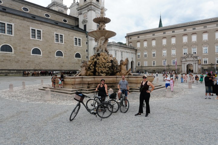 Three people with bicycles standing near a large fountain in a historic plaza.