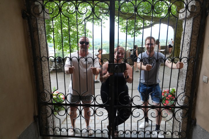 Three people smiling behind an ornate metal gate with a scenic view.