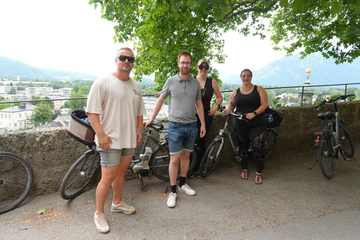 Four people with bicycles pose under a tree with a scenic view in the background.