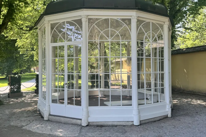White octagonal gazebo with glass windows set in a garden with green trees.