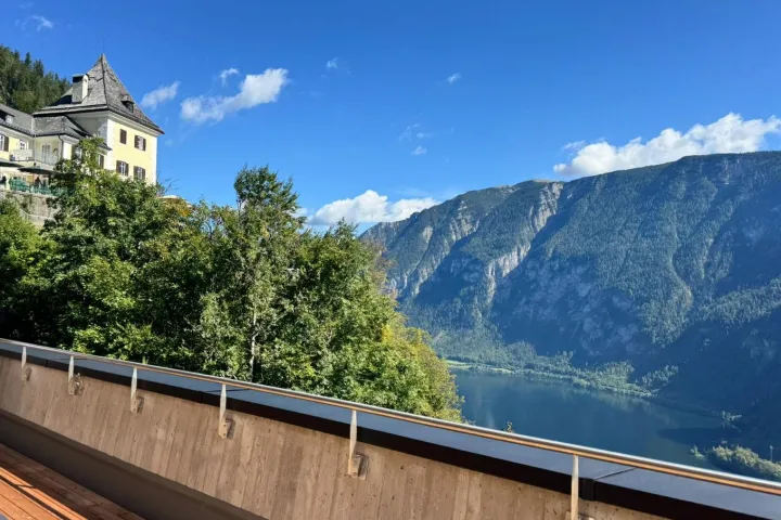 Mountain view with a building, trees, and lake under a clear blue sky.
