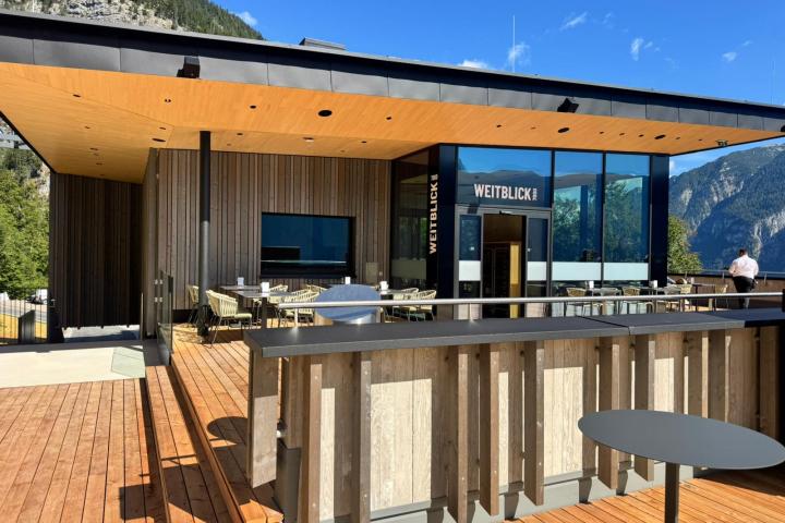 Modern building with large windows and wooden terrace, set against a mountain backdrop.