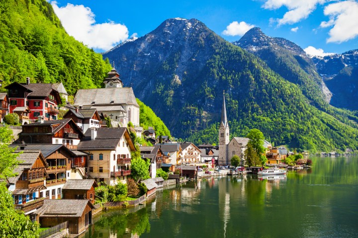 Scenic lakeside town with church steeple and mountain backdrop, surrounded by lush greenery and clear sky.