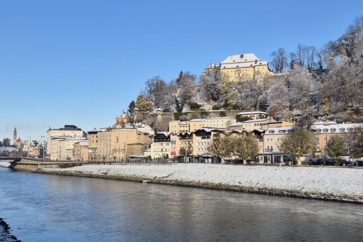 Snow-covered town by a river with hills and clear blue sky in the background.