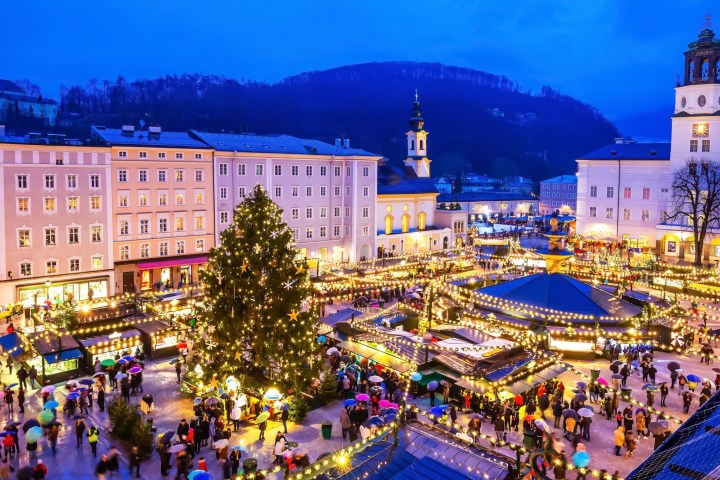 A bustling Christmas market with bright lights, a large tree, and people holding umbrellas at dusk.