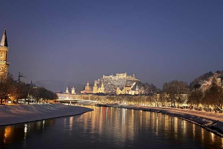 Night view of a snowy riverside city with illuminated castle and church.