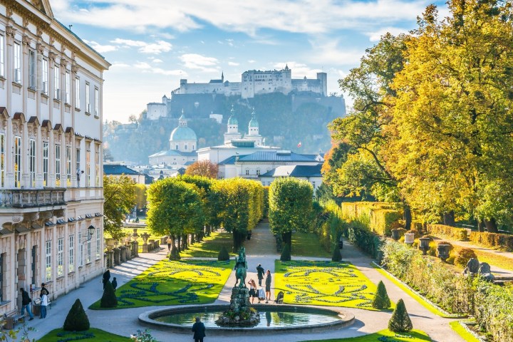 Ornate garden with fountain, path, and people, overlooked by a castle and autumn trees.
