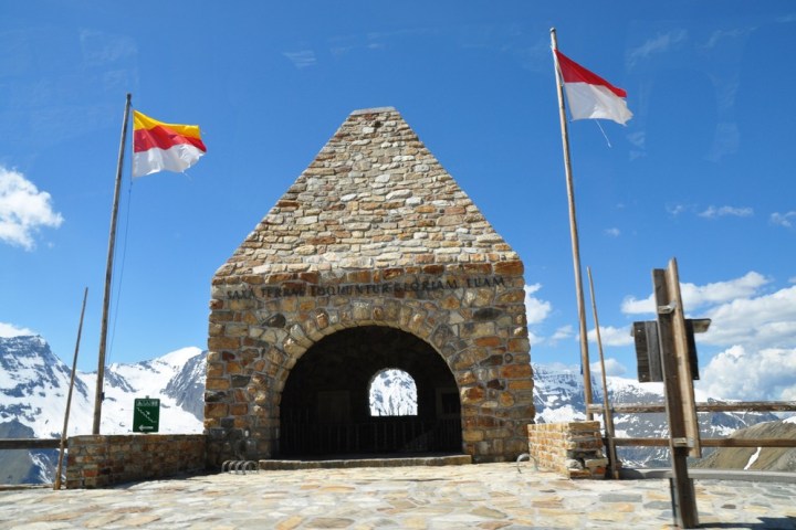Stone arch structure with flags on both sides, snowy mountains and blue sky in background.