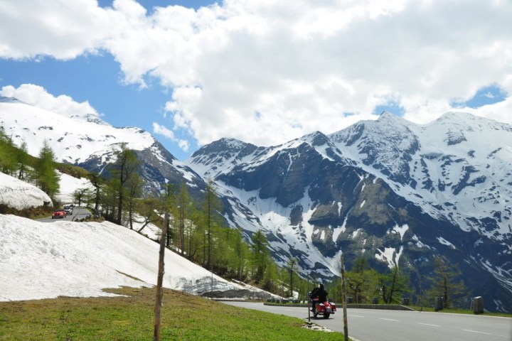 Snowy mountains with a winding road and motorcyclist, under a partly cloudy sky.