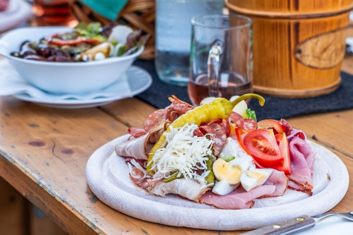 Plate of assorted cold cuts, sliced tomatoes, and eggs on a wooden table with salad bowls in the background.