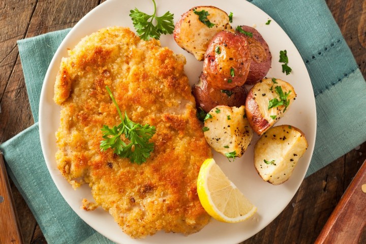 Breaded cutlet with roasted potatoes, garnished with herbs and lemon slice, on a plate with cutlery.