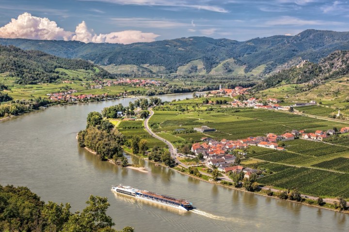 Scenic river with cruise ship, surrounded by vineyards and hills under a blue sky.
