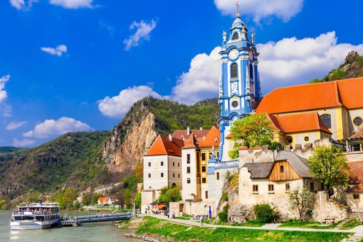 Colorful buildings and a blue church tower by a river, with hills and blue sky in the background.