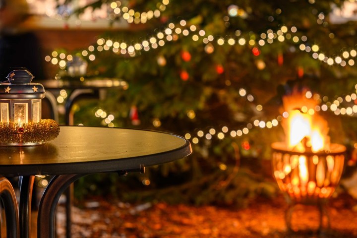 Lantern on table near Christmas tree with lights and fire pit in background.