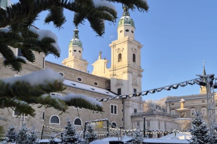 Snow-covered evergreen branches framing a historic building with towers on a clear blue sky.