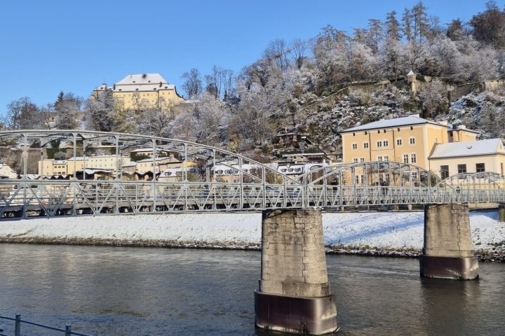 Snow-covered bridge and buildings beside a river with trees on a hill.