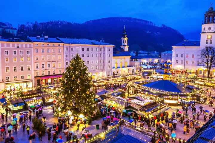 Festive Christmas market at night with lights, a large tree, and a crowd under umbrellas in a city square.