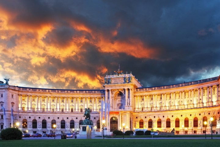 Illuminated baroque palace with dramatic sunset sky and dark clouds, featuring statues and green landscape in front.