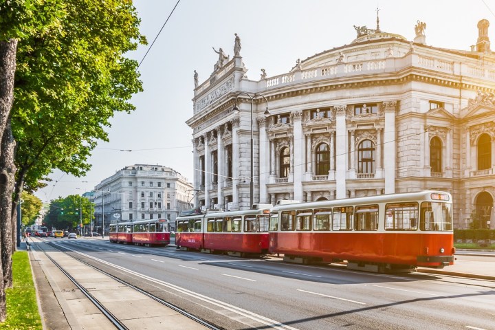 Red tram passing a historic building on a tree-lined street.
