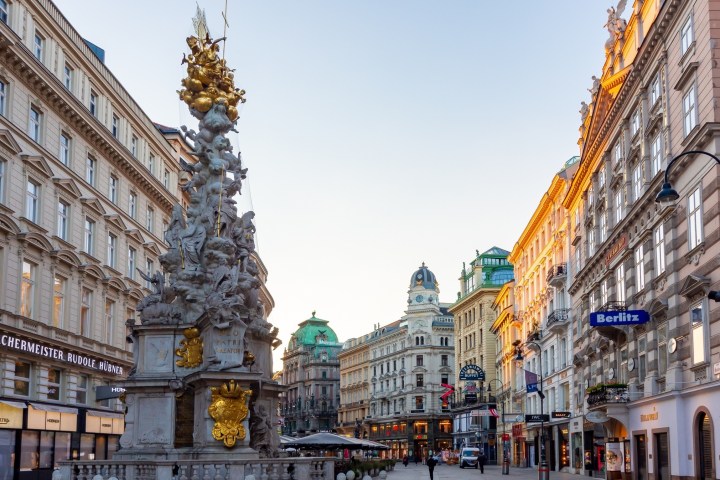 Historic street in Vienna with ornate buildings and a baroque monument at sunset.