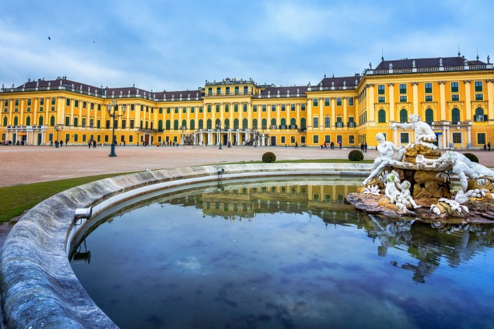 Majestic yellow palace with statues reflected in a circular fountain under a cloudy sky.