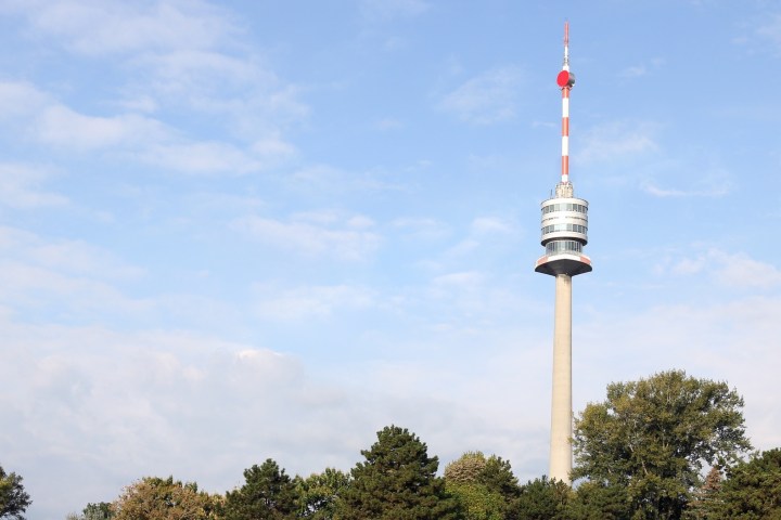 Tall communication tower with red and white antenna above trees under blue sky.