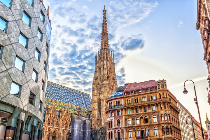 Gothic cathedral spire surrounded by modern and historic buildings under a partly cloudy sky.