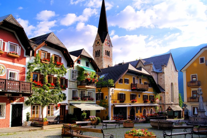 Colorful European village square with church spire, cobblestone paving, and flower-decorated buildings.