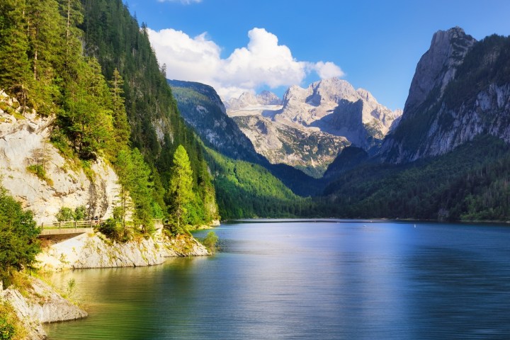 Scenic view of a mountain lake with lush forests and distant peaks under a blue sky with clouds.