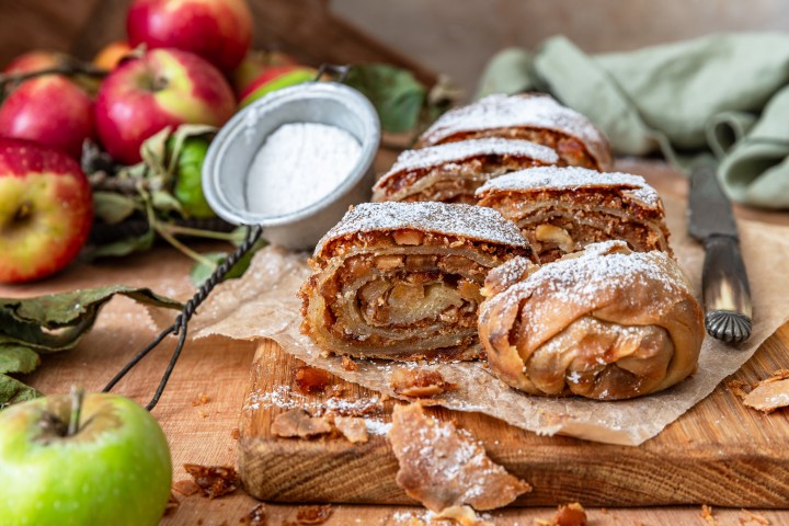 Sliced apple strudel with powdered sugar, apples, and a knife on a wooden board.
