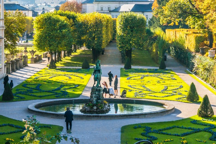 Formal garden with fountain, hedges, and people walking on pathways in front of an ornate building.