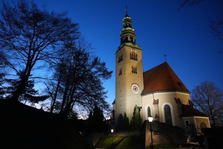 Church with tall tower at dusk, silhouetted trees, and illuminated clock.