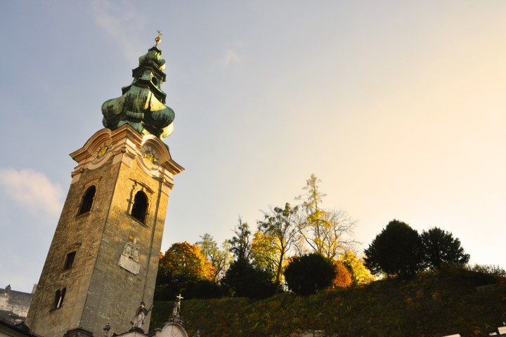 Historic tower with ornate spire against a sunset sky, surrounded by autumn trees.