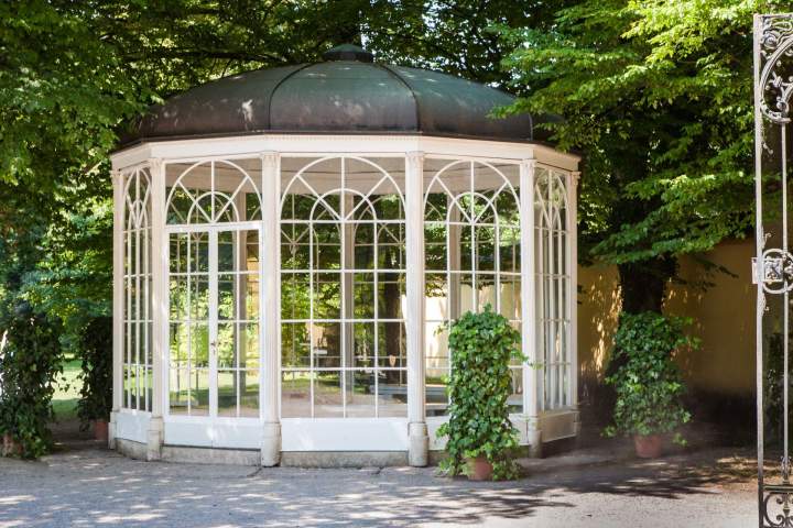 White gazebo with large windows surrounded by trees in a garden.