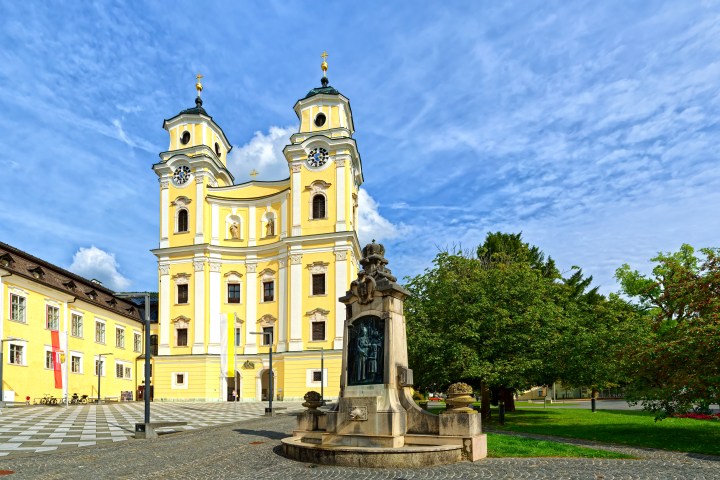 Yellow baroque church with two towers and a statue, surrounded by trees and a clear sky.