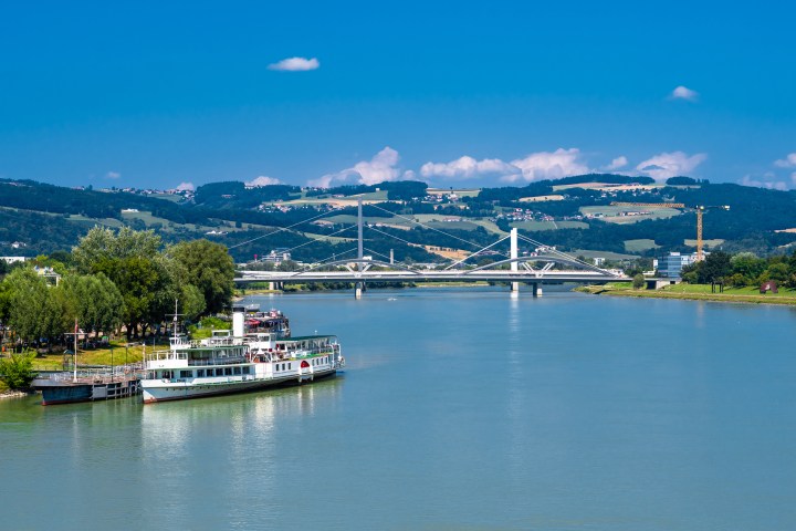 River with a boat, bridge, and hilly landscape under a clear blue sky.