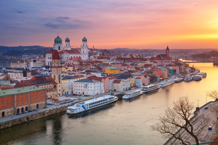 Sunset over a European city with colorful buildings and riverboats on a calm river.