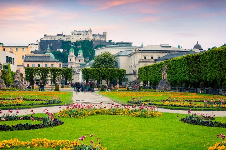 Garden with colorful flowers, path, trees, and historic buildings in the background under a pinkish sky.