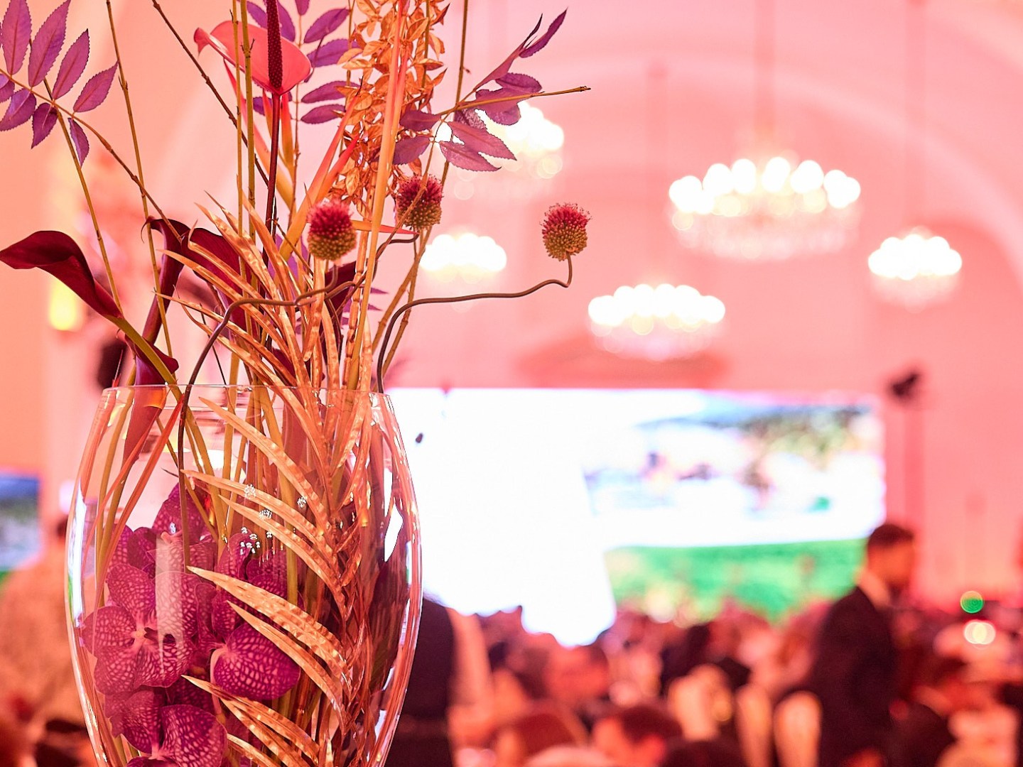 Elegant vase with flowers at event, blurred crowd and chandeliers.