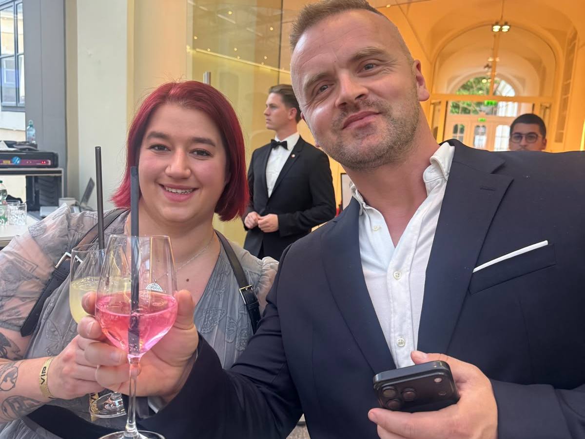 Two people at an event, holding drinks and smiling, with a formal dressed person in the background.
