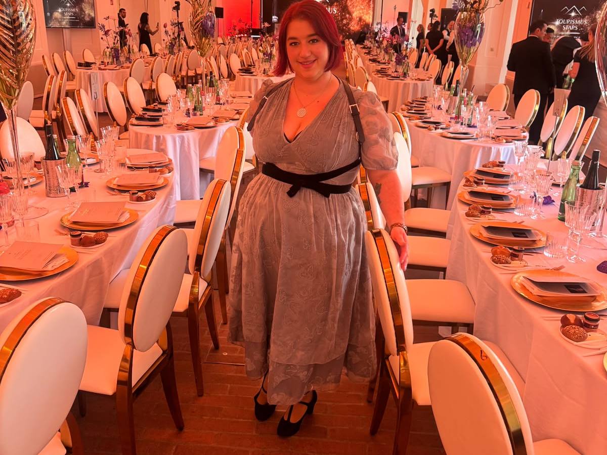 Person in a gray dress stands in a grand dining hall with white tables and chandeliers.