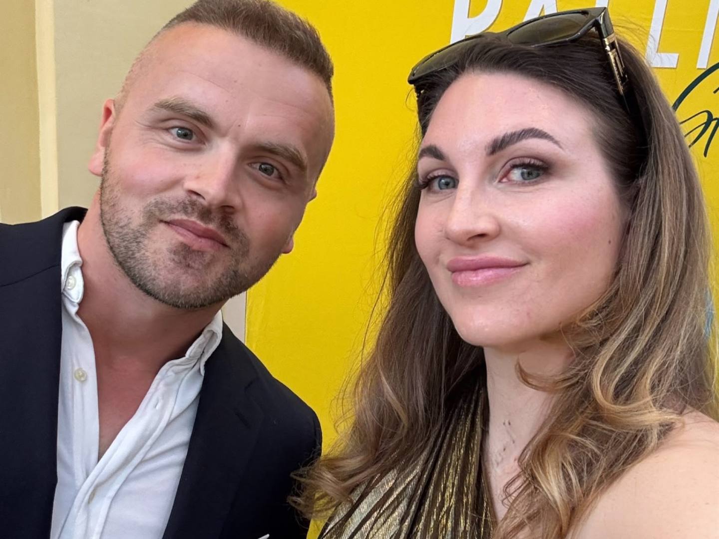 Two people smiling, one in a suit, one in a gold dress, in front of a yellow sign.
